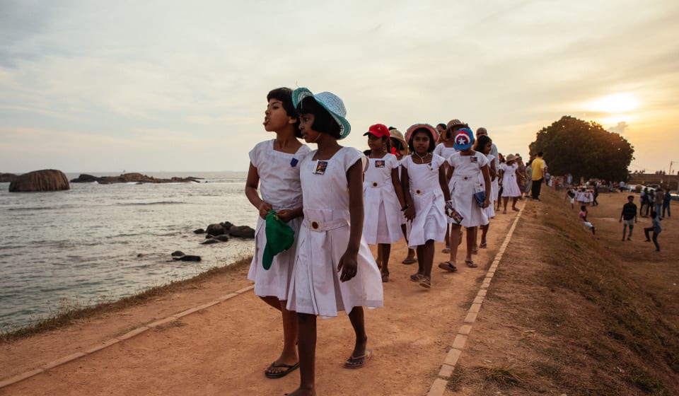 A group of kids walking in a row in pairs of 2 next to the water in Sri Lanka