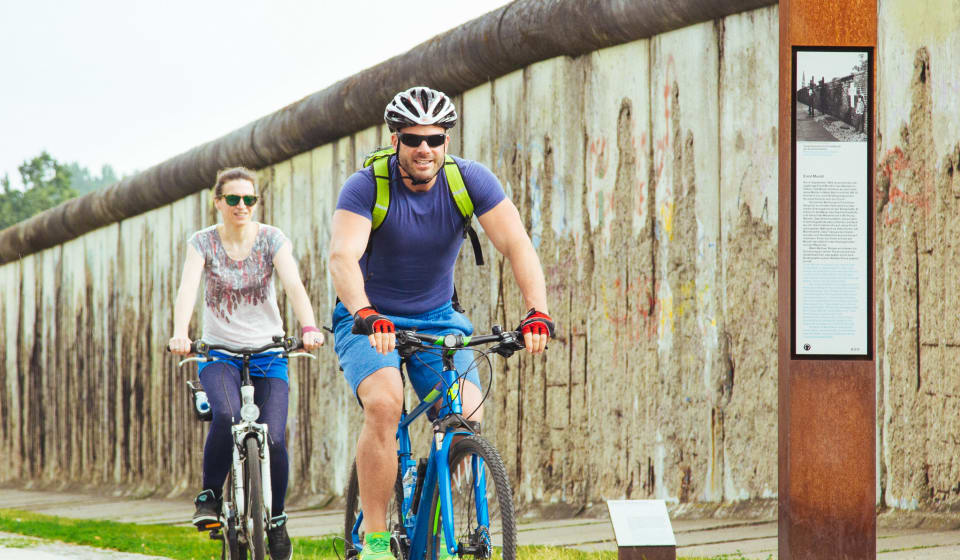 A local guide cycling with a tourist next to a the Berlin Wall