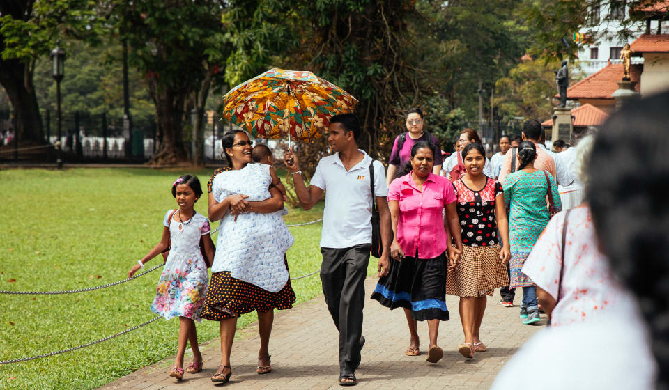 A group of locals walking in a park in Sri Lanka. One guy in the group has an umbrella above his and a lady's head