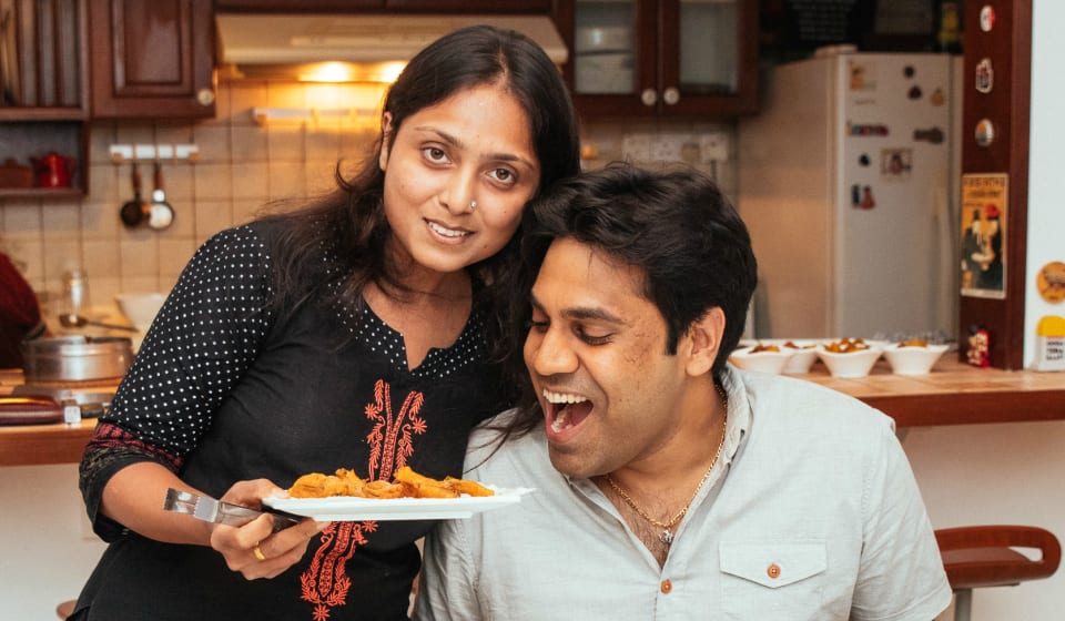 A lady showing a plate of Singaporean food to a guy. The guy is attempting to take a bit of the food