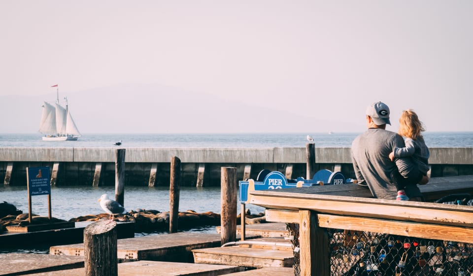 A man with a child on his arms looking over a pier in San Franciso to a sail boat
