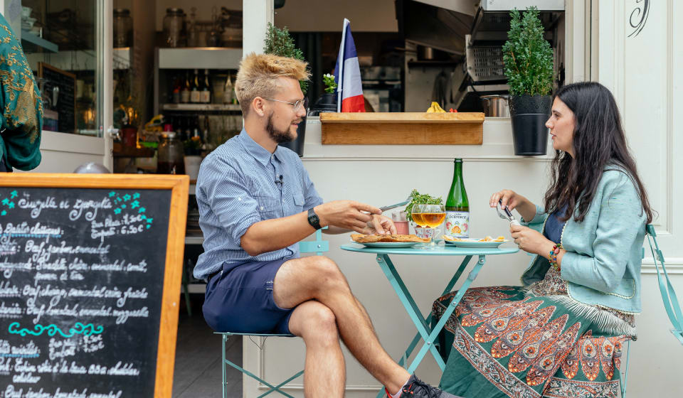 Two people eating French crepes at a table outside with a French flag in the background