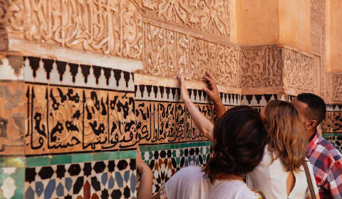 A guide showing a wall with Arabic scripture on the a wall with colorful tiles in Marrakech to two tourists