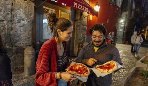A local guide and a tourist eating a pizza on the streets of Naples