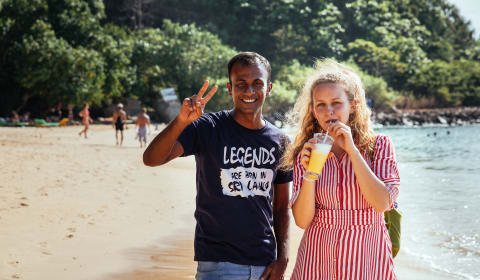 A man and woman on the beach of Mount Lavinia. The man is making a victory sign with his fingers and the woman is drinking a King Coconut