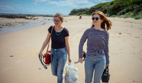 Two ladies walking on a beach with bags